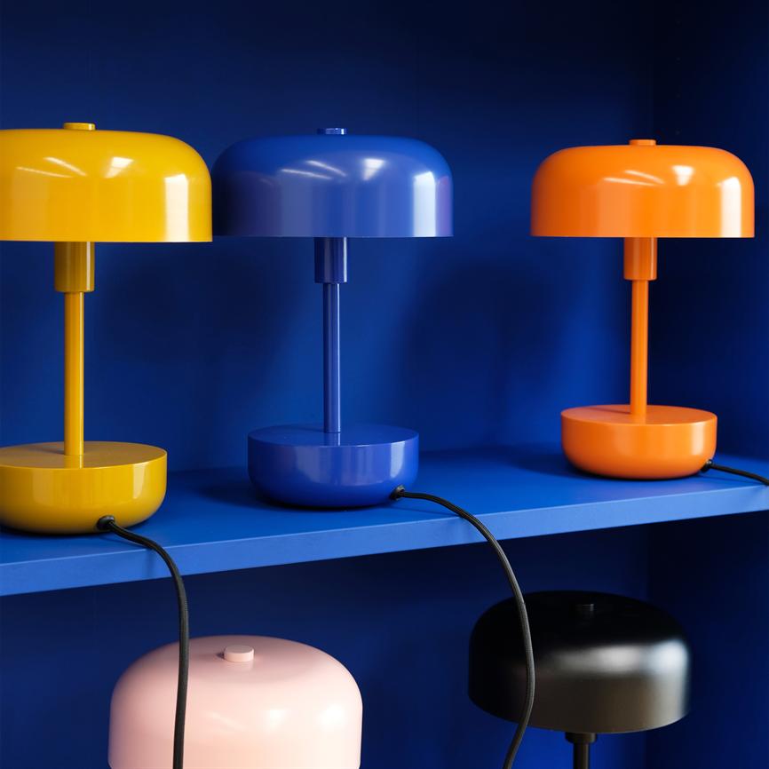 Drei glatte, runde Tischlampen in Gelb, Blau und Orange stehen nebeneinander auf einem blauen Regal, verbunden durch schwarze Kabel vor einem tiefblauen Hintergrund.