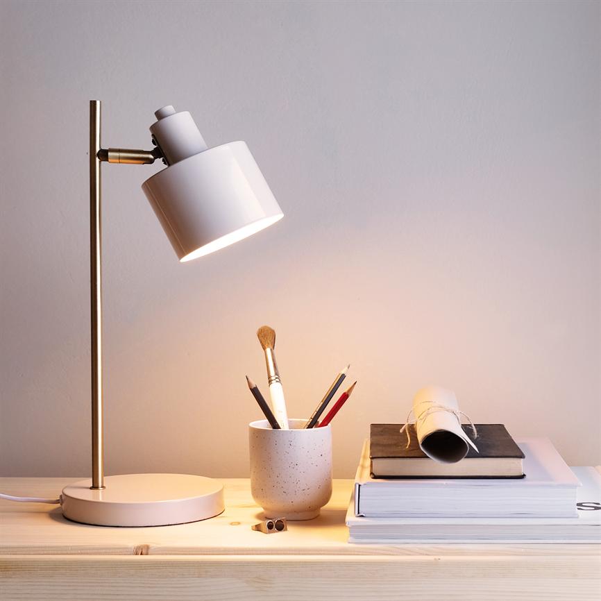 Eine weiße Tischlampe scheint auf eine Porzellantasse mit Stiften und Pinsel, die auf einem hellen Holztisch neben einem Stapel Bücher und einem aufgerollten Papier an einer hellen Wand steht.