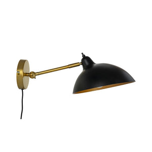 Væglampe med sort kuppelskærm og guldfarvet metalarm, monteret på et rundt vægbeslag; armen er udstrakt og skærmen vippet nedad mod en hvid, neutral baggrund.