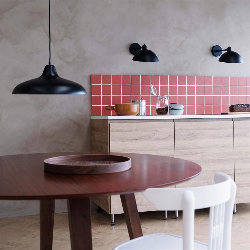 Vorne steht ein runder Holztisch mit einem runden Holztablett auf der Oberfläche; Dahinter befindet sich eine helle Holzküche mit rosa Fliesen, schwarzen Lampen an der Wand und einer hängenden schwarzen Pendellampe.