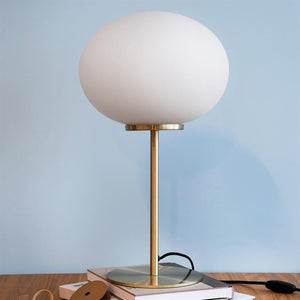 Tischlampe mit großer weißer Glaskuppel steht auf einem Messingstab, platziert auf einem Bücherstapel auf einem Holztisch, vor einer hellblauen Wand.