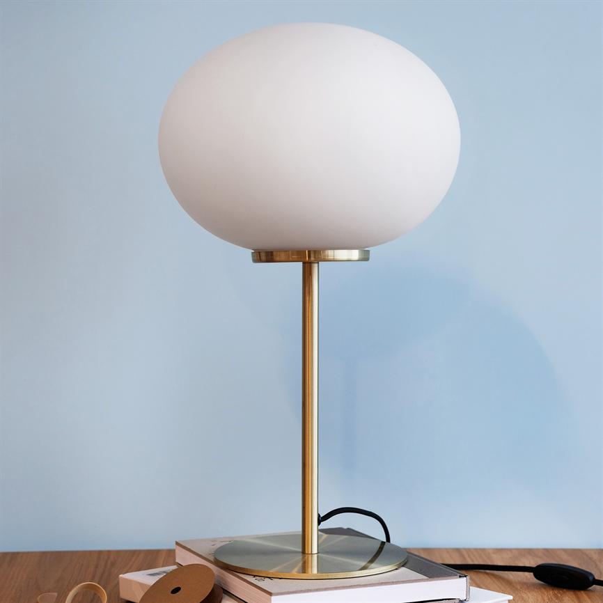 Tischlampe mit großer weißer Glaskuppel steht auf einem Messingstab, platziert auf einem Bücherstapel auf einem Holztisch, vor einer hellblauen Wand.