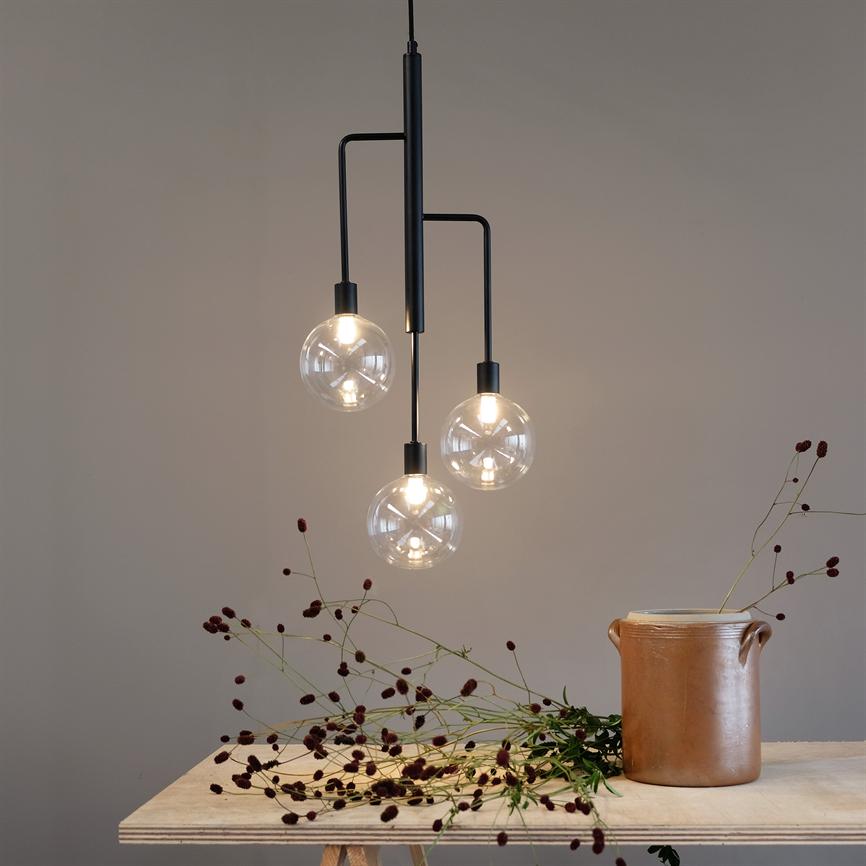 Ein schwarzer pendel mit drei beleuchteten Glaskugeln hängen über einem hellen Tisch mit einem braunen Keramikgefäß und verstreuten getrockneten dunkelroten Blumen vor einer beigen Wand.