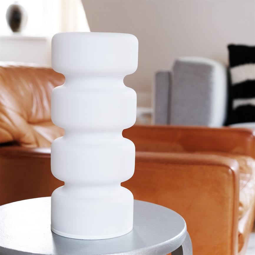 Weiße skulpturale Tischlampe steht auf einem runden silberfarbenen Tisch vor einem braunen Ledersofa und einer grauen Sofaecke in einem hellen, modernen Wohnzimmer.