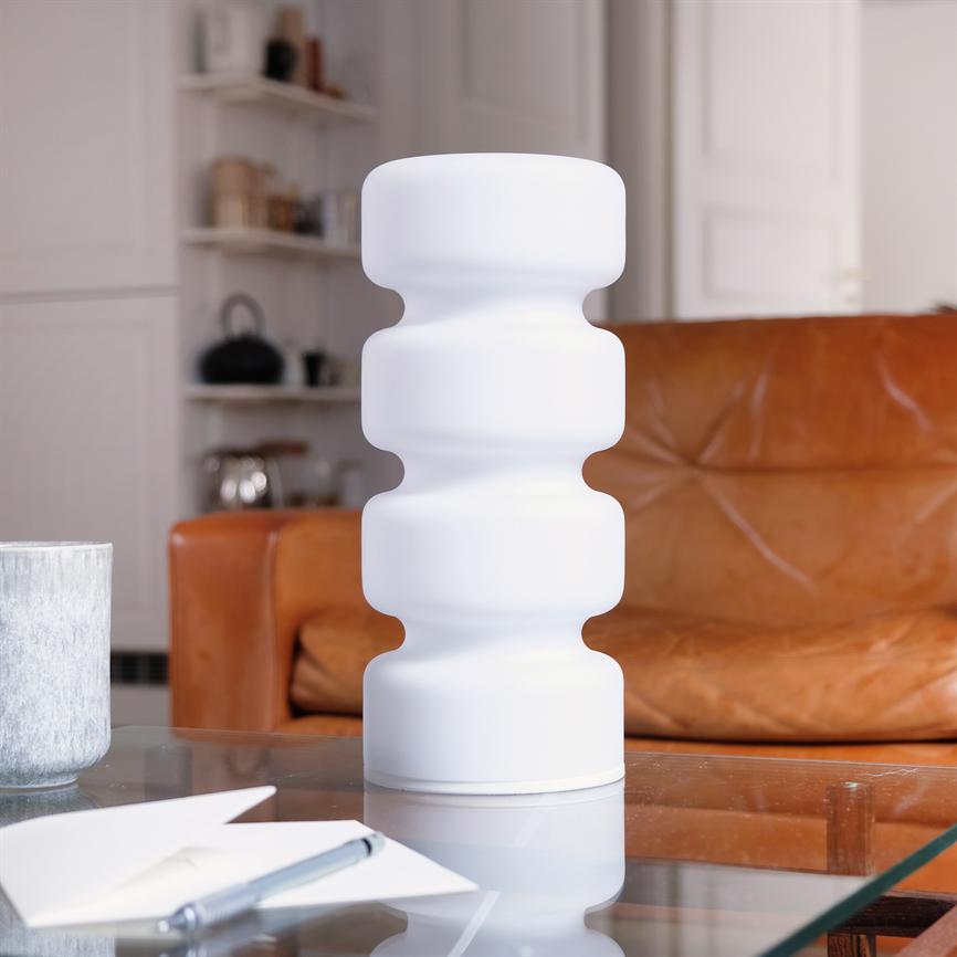 Weiße, gewellte Tischleuchte steht auf einem Glastisch; umgeben von Notizblock und Stift im Vordergrund, vor einem braunen Ledersofa und Regalen in einem gemütlichen Wohnzimmer.