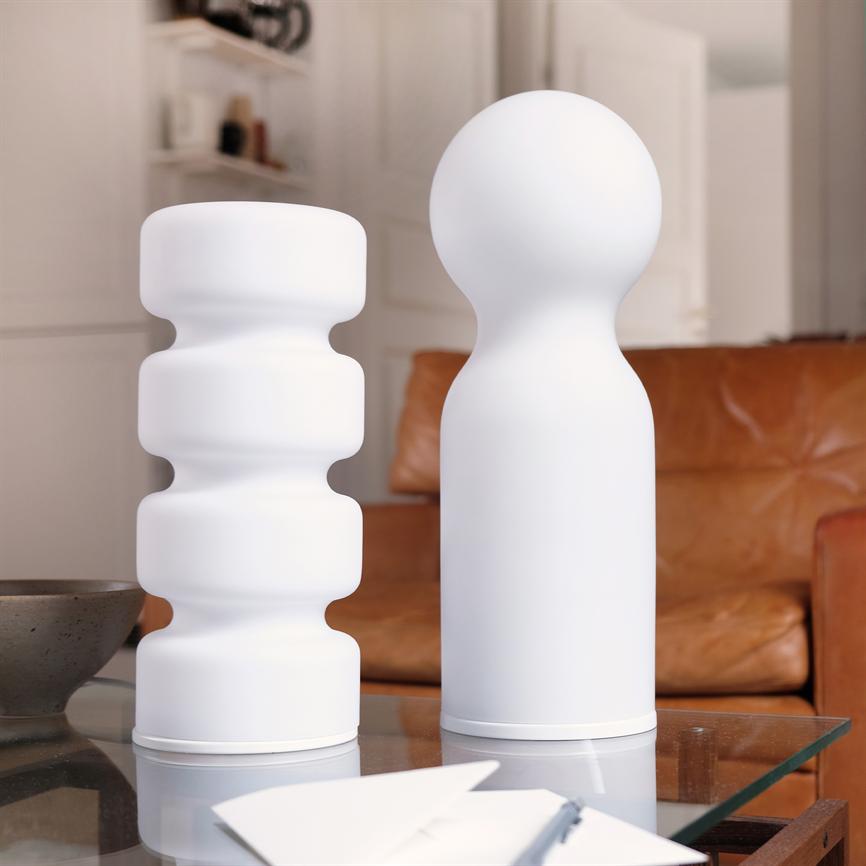 Zwei weiße, skulpturale Vasen stehen nebeneinander auf einem Couchtisch aus Glas; Eine davon ist gewellt, die andere hat eine runde Oberseite, vor einem braunen Ledersofa in einem hellen Wohnzimmer.