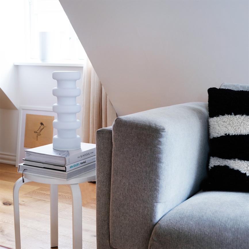 Eine weiße geriffelte Vase ruht auf einem Stapel Bücher auf einem kleinen weißen Beistelltisch; neben einem grauen Sofa mit schwarz-weißem Kissen unter einer schrägen Deckenwand und Licht aus einem Fenster. 
 
 Transkription des sichtbaren Textes: „THE KINFOLK HOME“, „ARA GÜLER: Istanbul“