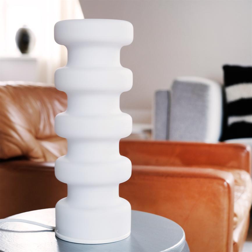 Weiße, skulpturale Tischleuchte steht auf einem runden Beistelltisch; Die wellenförmige Zylinderform bildet einen Kontrast zu braunen Ledersesseln und grauen Polstermöbeln in einem hellen, modernen Wohnzimmerinterieur.