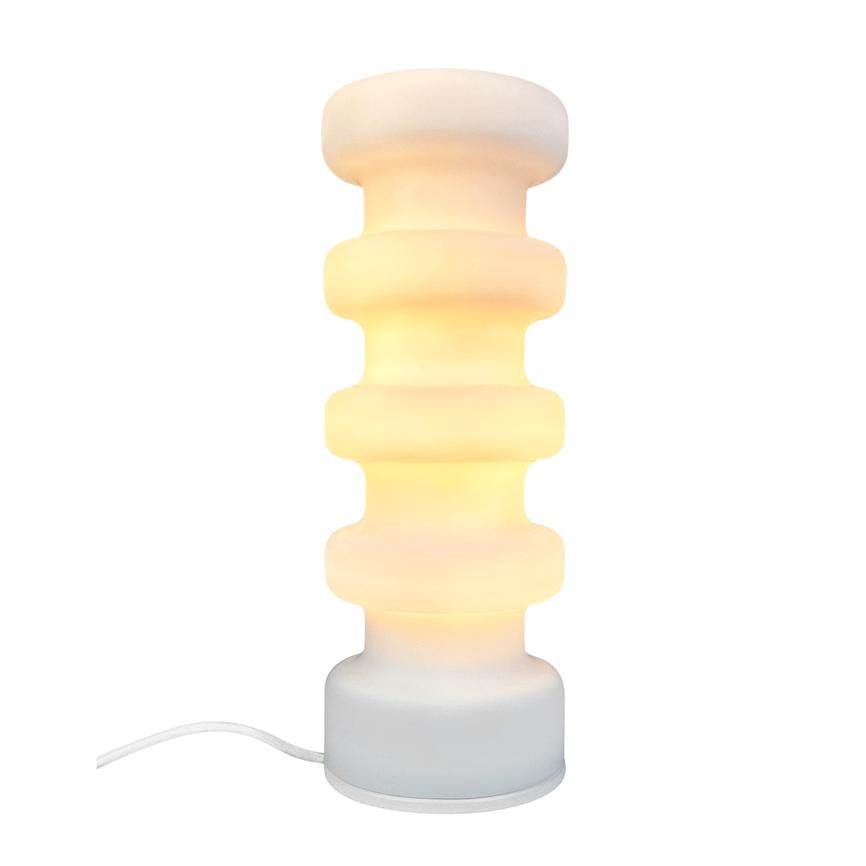 Bordlampe med stabel af brede, afrundede skiver lyser varmt; den står på en rund fod med hvid ledning synlig mod en ensartet hvid baggrund, i et minimalistisk miljø.