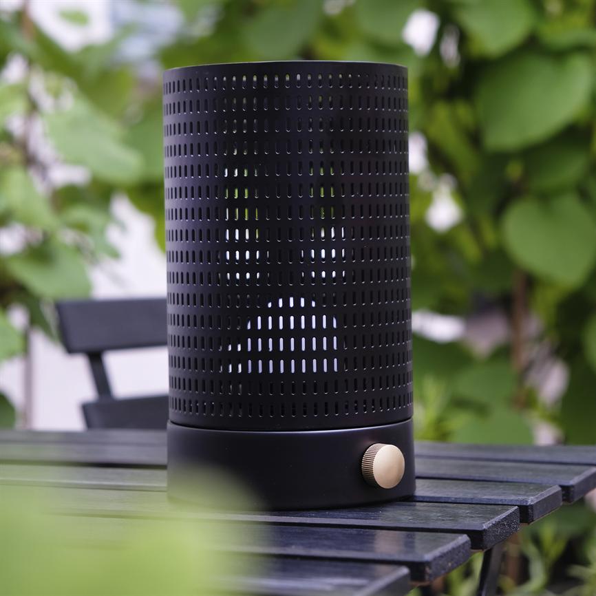 Schwarzer, perforierter zylindrischer Lautsprecher mit goldenem Zifferblatt steht auf einem dunklen Holztisch; Außenumgebung mit verschwommenen grünen Pflanzen und einem schwarzen Gartenstuhl im Hintergrund.