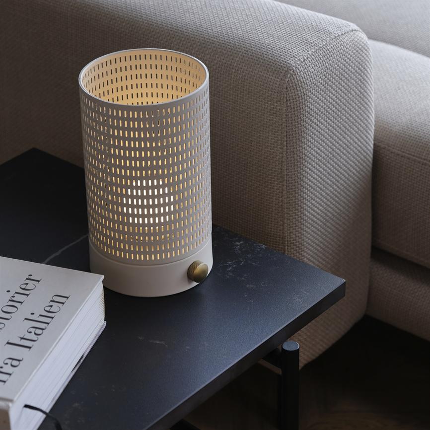 Eine zylindrische, perforierte Tischlampe strahlt ein warmes, gut beleuchtetes Licht aus und steht auf einem dunklen Beistelltisch neben einem beigen Sofa neben einem Buch mit der Aufschrift „Geschichten aus Italien“.