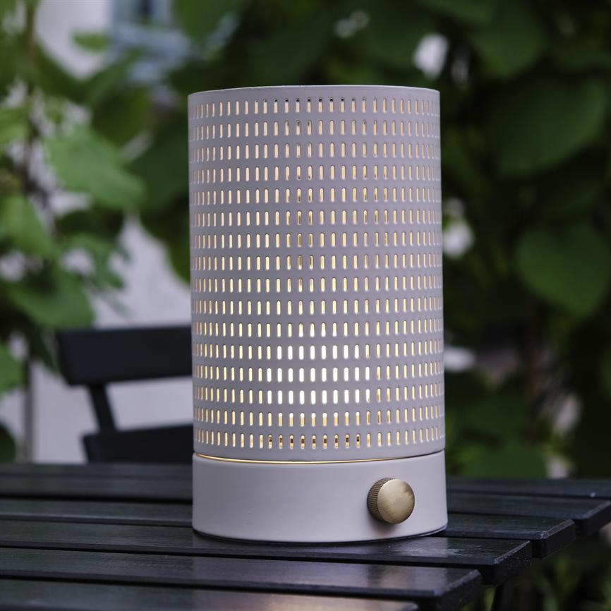 Eine zylindrische Lampe aus perforiertem Metall leuchtet warm durch rechteckige Löcher; Messingknopf an der Basis, platziert auf einem schwarzen Lattentisch im Freien mit verschwommenem grünem Blatthintergrund.