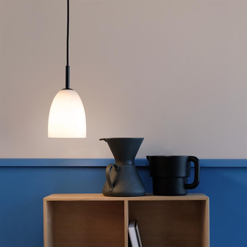 Eine weiße, kuppelförmige Hängelampe leuchtet sanft über einem kleinen Holzregal, auf dem vor einer blau-beigen Wand eine schwarze, trichterförmige Kaffeekanne und ein schwarzer Becher stehen.