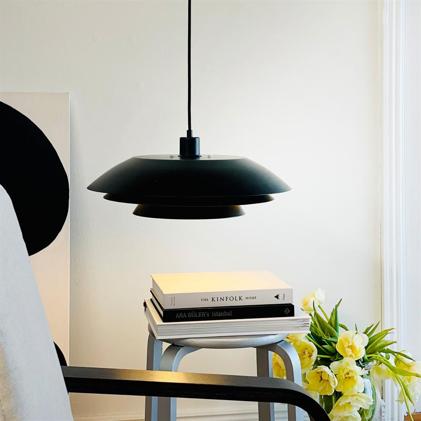 Schwarz pendel-Lampe hängt über einem Stapel Bücher und spendet sanftes Licht; Die Bücher liegen auf einem kleinen Hocker neben einem Stuhl und einem Strauß gelber Tulpen in einem hellen Raum.
 
 Text im Bild: THE KINFOLK HOME; ARA GÜLERS Istanbul