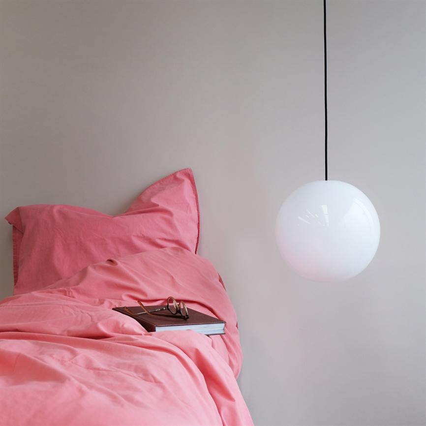 Ein rosa Bettset liegt zusammengerollt auf einem Bett, darauf liegen ein Buch und eine Brille; ein runder, weißer pendel hängt vor einer neutralen Wand und bildet einen schlichten Hintergrund.
