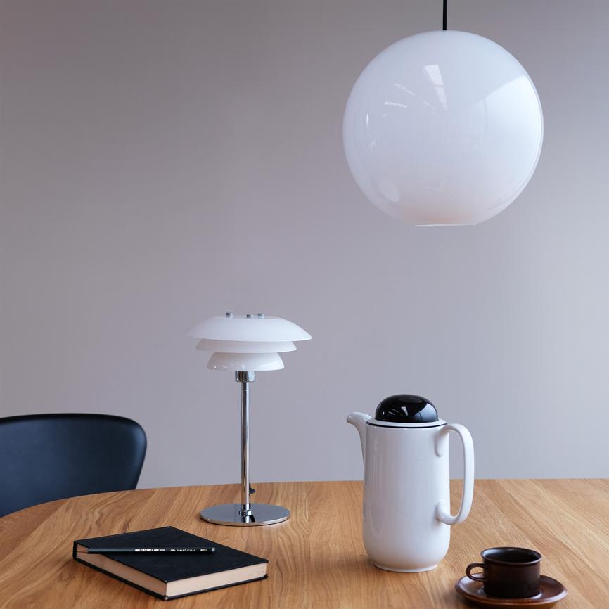 Weiße kugelförmige Deckenleuchte hängt über einem hellen Holztisch; Auf dem Tisch stehen eine weiße Thermoskanne, eine kleine dunkle Kaffeetasse, ein Notizbuch und eine moderne Tischlampe in einem minimalistischen Raum.