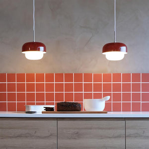 Zwei rote Pendelleuchten strahlen auf einen Küchentisch, auf dem auf einem Schneidebrett neben einer weißen Schüssel ein Laib Roggenbrot liegt; Die Rückwand ist mit roten quadratischen Fliesen bedeckt.