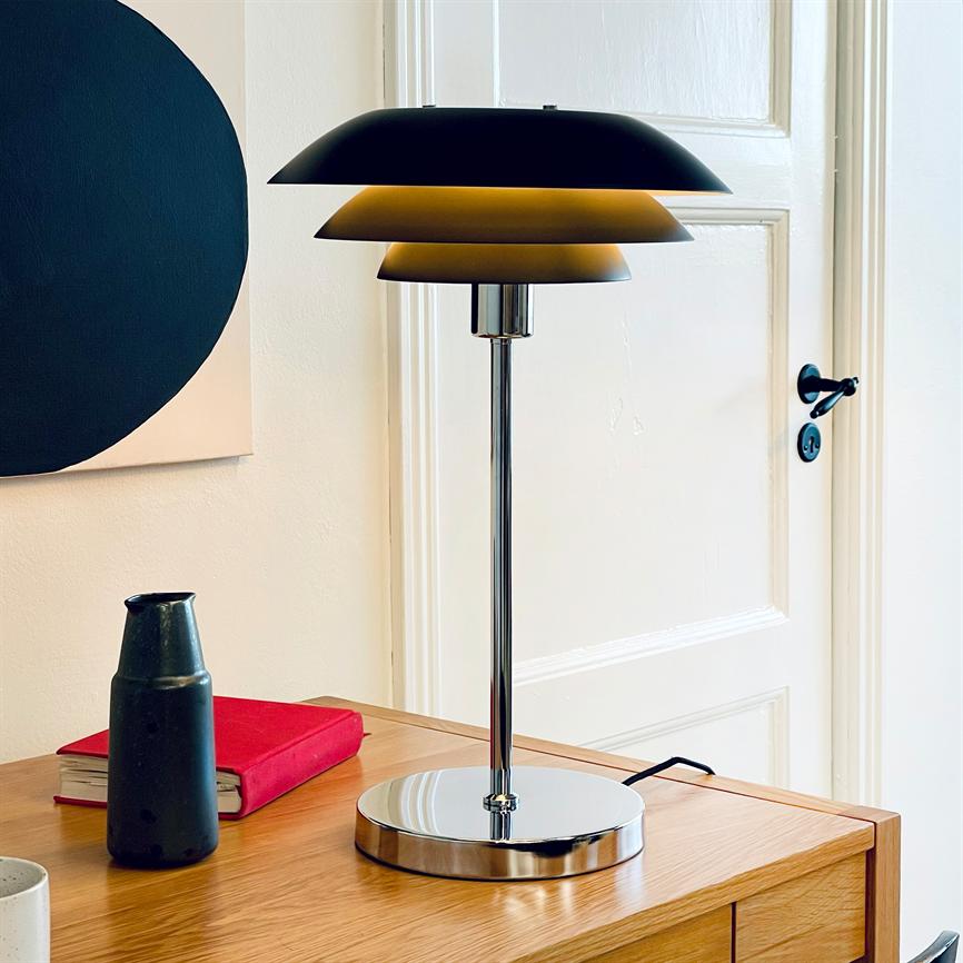 Die verchromte Tischlampe mit drei Schirmen leuchtet warm und steht auf einem Holzschreibtisch neben einer weißen Tür, flankiert von einem roten Buch, einem schwarzen Krug und abstrakter Wandkunst.