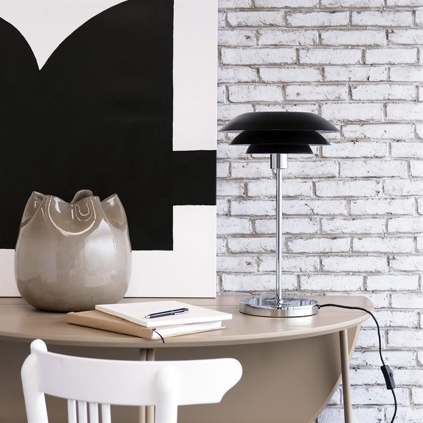 In der Mitte eines runden beigen Schreibtisches steht eine schwarze, mehrschichtige Tischlampe; daneben liegen Notizbücher und Stift, eine graue Keramikvase und ein schwarz-weißes abstraktes Gemälde vor einer weißen Backsteinwand.
