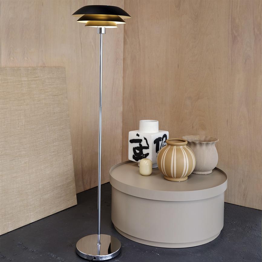 Gulvlampe står ved et rundt beige bord; lampens sorte og guldskærm kaster varmt lys over tre keramikkrukker og en firkantet hvid vase foran lyst træpanel. 
 Tekst på vase: utydelige sorte penselstrøg/kaligrafi, ikke entydigt aflæseligt.