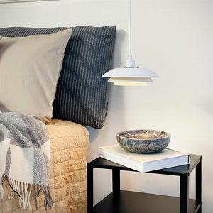 Eine weiße Pendelleuchte leuchtet schwach über einem schwarzen Nachttisch mit einem Buch und einer Marmorschale; Daneben steht ein Bett mit beiger Tagesdecke, grau gestreiftem Kopfteil und karierter Decke.