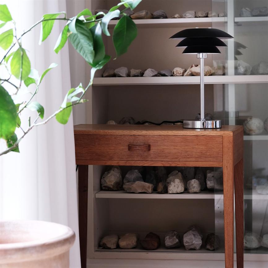 Ein kleiner Holztisch mit Schublade steht vor einem Bücherregal; Auf der rechten Seite des Tisches steht eine schwarze, moderne Tischlampe, umgeben von Steinregalen und einer grünen Pflanze am Fenster.