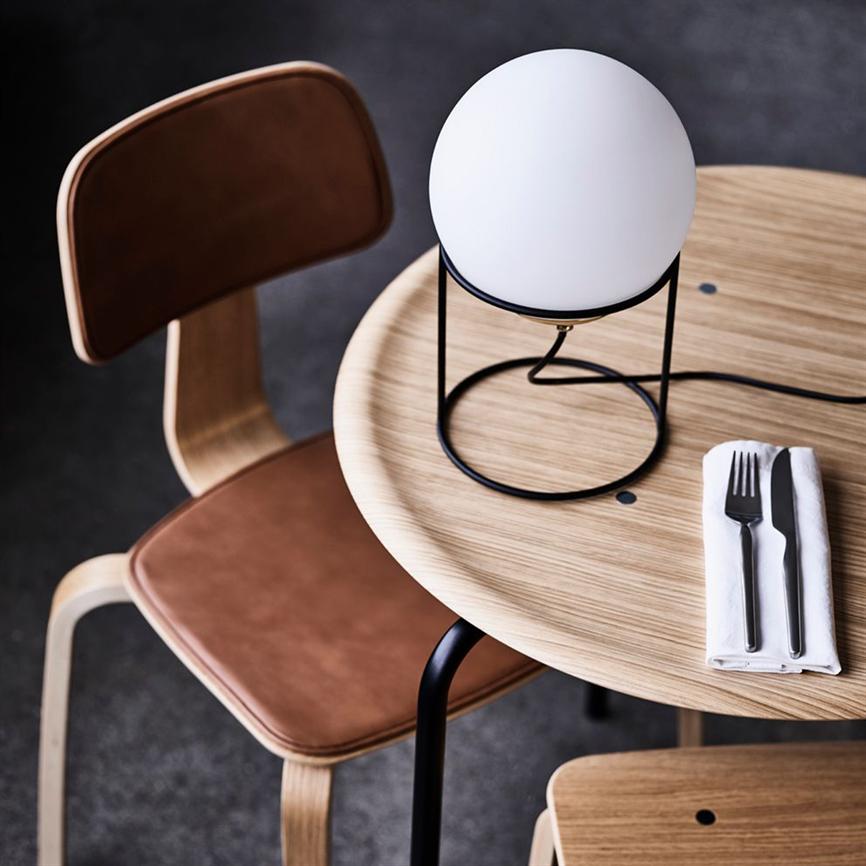 Runde Tischleuchte steht in einem schwarzen Metallständer auf einem runden hellen Holztisch; Kabel verläuft über den Tisch. Am Rand liegt Besteck in einer weißen Serviette. Ein brauner Ledersessel fährt herein.