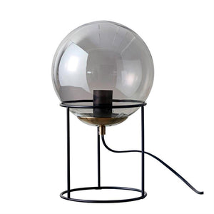 Runde Kuppellampe aus Rauchglas, die auf einem dreibeinigen schwarzen Metallständer ruht; Steckdose und schwarzes Kabel sichtbar, platziert vor einem einheitlichen weißen Studiohintergrund.