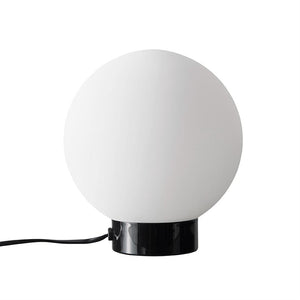 Kuglelampe: en stor hvid glaskugle sidder på en blank sort cylindrisk fod, med en sort ledning som løber ud til siden, placeret mod en neutral hvid baggrund.