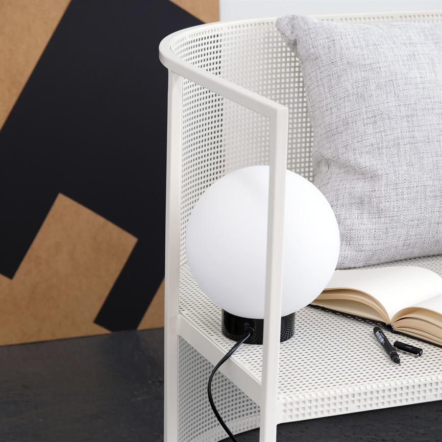 Auf einem weißen, perforierten Regal steht eine runde weiße Tischlampe; daneben liegen ein aufgeschlagenes Notizbuch und ein Stift, im Hintergrund ein hellgraues Kissen und ein schwarzbraunes Poster.