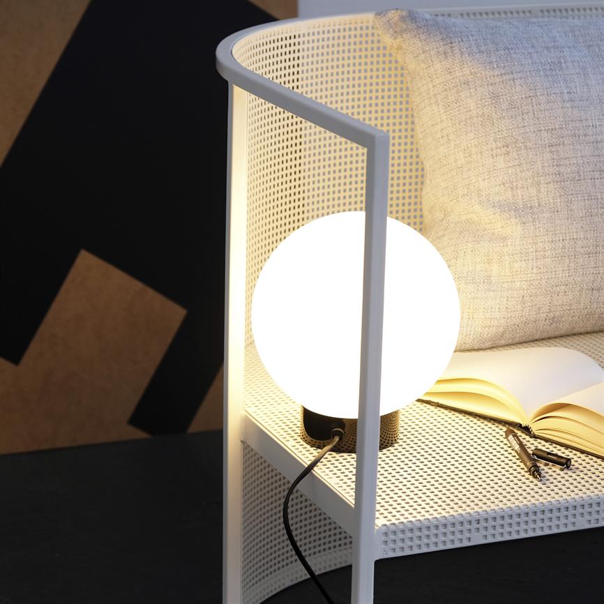 Die runde Tischlampe erstrahlt warm auf einem weißen, perforierten Beistelltisch neben einem hellgrauen Kissen und einem aufgeschlagenen Buch mit Stift in einer modernen, dezenten Ecke.