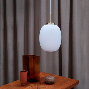 Ein weißes Oval pendel hängt über einem Holztisch; Auf dem Tisch stehen eine hohe braune Glasvase, eine kleine Terrakotta-Tasse und ein Bindfadenschlüssel mit loser Schnur. Der Hintergrund sind graue Vorhänge.