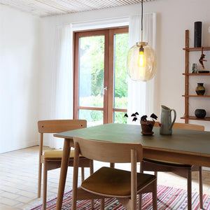 Der Esstisch mit vier Holzstühlen steht unter einer großen Hängelampe aus Glas; auf dem Tisch stehen ein Krug und eine kleine Topfpflanze; Große Fenster öffnen sich zu einem grünen Gartenteil in einem hellen Raum.