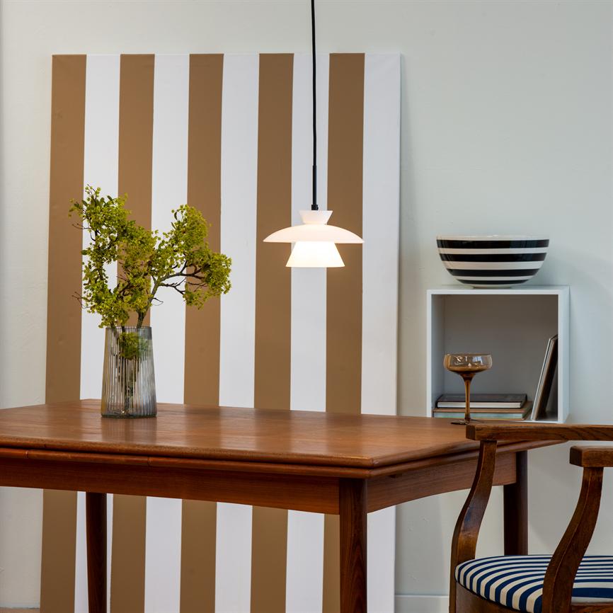 Auf einem Holztisch steht eine klare Glasvase mit grünen Zweigen, während eine weiße Pendelleuchte hängt und leuchtet; Hinter dem Tisch befindet sich eine vertikal braun-weiß gestreifte Platte, ein Regal mit einer Schüssel und einem Stuhl.