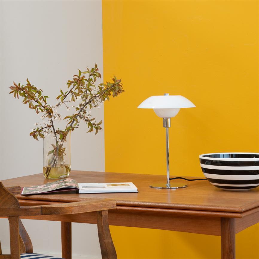 Auf einem Holztisch steht eine weiße Tischlampe; links eine Glasvase mit Blumen, vorne ein aufgeschlagenes Buch und rechts eine schwarz-weiß gestreifte Schale; die Rückwand ist in Weiß und Gelb unterteilt.