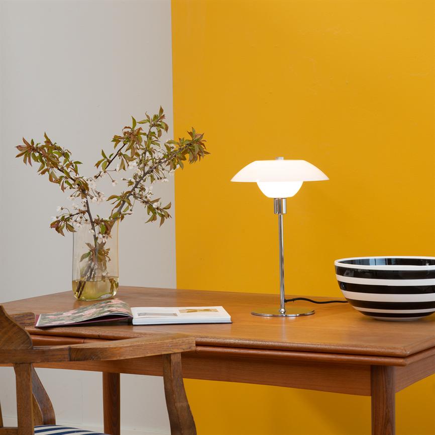 Tischlampe leuchtet sanft über einem Holztisch; links Glasvase mit Blumenzweigen und aufgeschlagenem Buch, rechts schwarz-weiß gestreifte Schale; Wand im Hintergrund in Weiß und Gelb unterteilt.