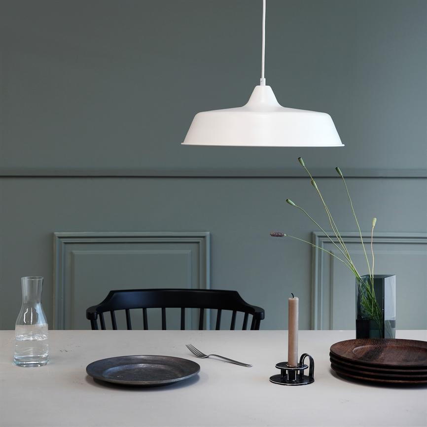 Weiß pendel hängt über einem gedeckten Tisch; dahinter ein schwarzer Stuhl, eine Glaskaraffe, ein Teller und eine Gabel, ein Kerzenhalter mit Kerzen und eine Vase mit hohen Blumen vor einer grünen Wand.