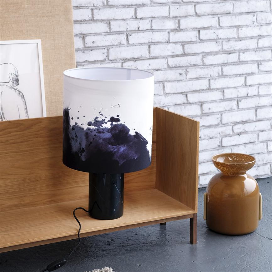 Auf einem niedrigen Holzregal steht eine Tischlampe mit weißem, mit dunkelblauen Aquarellspritzern gebeiztem Schirm; schwarzer Marmorsockel und Kordel, vor weißer Ziegelwand, mit brauner Keramikvase und gerahmter Skizze.