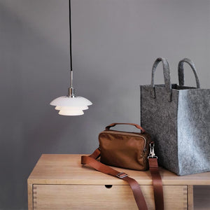 pendel hängt und wirft sanftes, warmes Licht über eine helle Holzkommode, auf der eine braune Umhängetasche mit einem Riemen über dem Rand liegt und daneben eine große graue Filztasche steht.