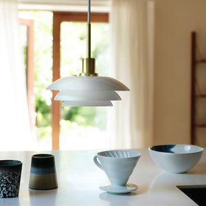 Eine weiße, mehrschichtige Pendelleuchte hängt und wirft sanftes Licht über einen Küchentisch mit Keramiktassen, einem Kaffeetrichter und einer Schüssel, vor großen Fenstern mit hellen Vorhängen und draußen im Grünen.