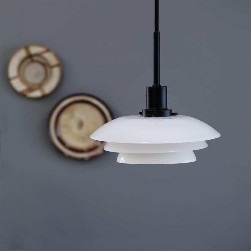 Eine weiß geschichtete Pendelleuchte hängt an einem schwarzen Kabel vor einer grauen Wand mit zwei verschwommenen runden dekorativen Holzpaneelen.