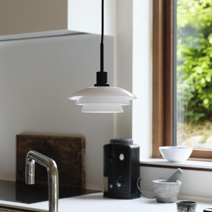 pendel mit geschichteten weißen Schirmen, die an einer schwarzen Kordel über dem Küchentisch hängen; Im Hintergrund sieht man einen verchromten Wasserhahn, eine Kaffeemaschine, einen Mörser und eine Schüssel am Fenster mit viel Grün draußen.