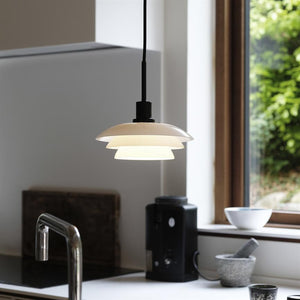 Über dem Küchentisch hängt eine weiße, abgestufte Pendelleuchte; Es wirft sanftes Licht auf die Spüle, die Kaffeemaschine und eine Schüssel am Fenster in einer hellen, modernen Küche.