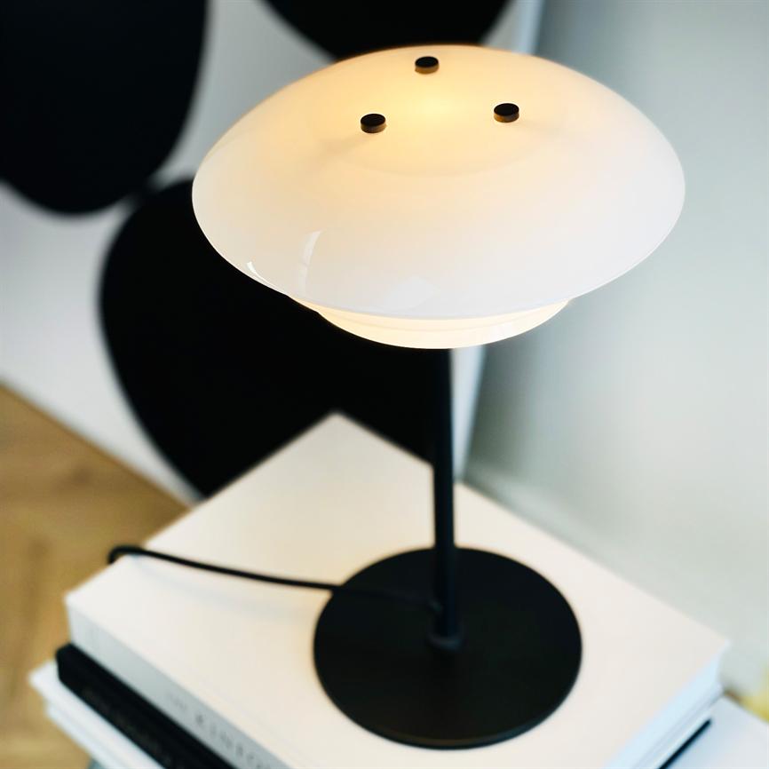 Eine schwarze Tischlampe mit weißem, schalenförmigem Schirm leuchtet sanft, platziert auf einem Stapel weißer Bücher auf einem Holzboden, mit einem undeutlichen schwarzen Hintergrund.