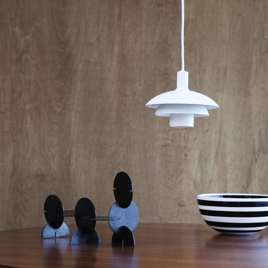 Über einem runden Holztisch hängt eine weiße Pendelleuchte, links auf dem Tisch steht ein schwarzes geometrisches Metallobjekt und rechts vor einer Holzwand eine schwarz-weiß gestreifte Schale.