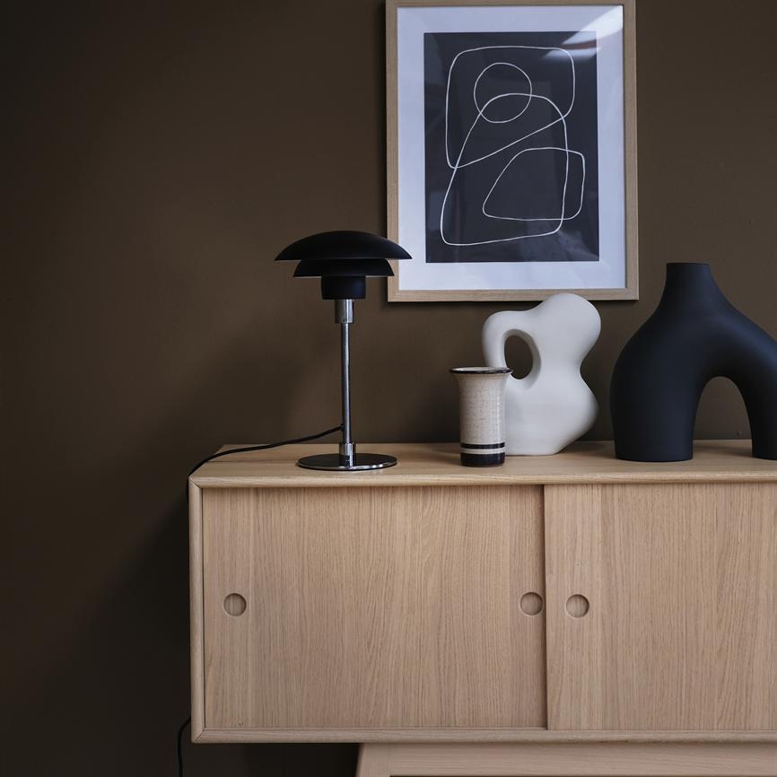 Auf einem Sideboard aus heller Eiche stehen eine schwarze Tischlampe, ein Keramikbecher und zwei skulpturale Vasen vor einer braunen Wand mit einem gerahmten abstrakten Schwarz-Weiß-Poster.