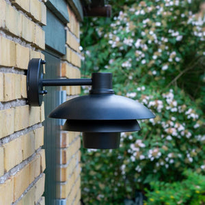 Schwarze Wandlampe an gelber Ziegelwand montiert, an einem kurzen Arm hängend; Lichtkuppel zeigt nach unten; Der Hintergrund zeigt einen grünen Garten mit Büschen und kleinen rosa Blüten.