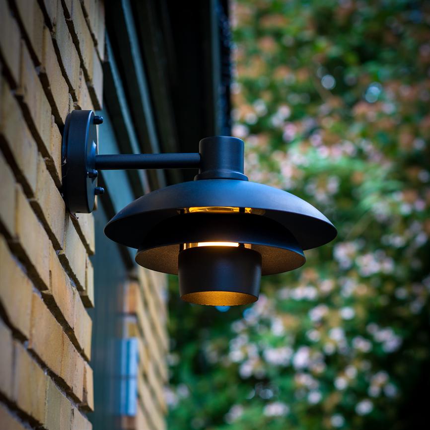 Eine schwarze, runde Wandleuchte strahlt warmes, nach unten gerichtetes Licht ab; Es ist an einer gelben Backsteinwand draußen montiert, mit verschwommenem grünem Blattwerk und Blumen im Hintergrund.