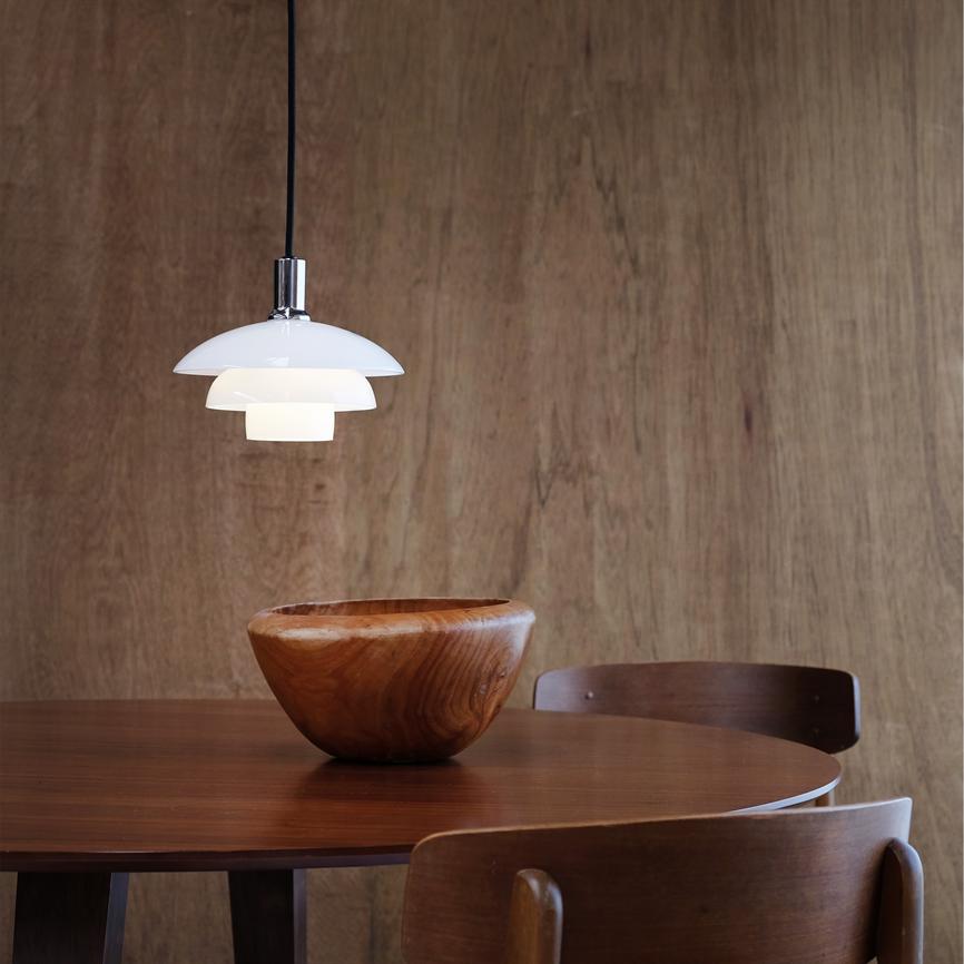 Weiß pendel hängt über einem runden dunklen Holztisch; In der Mitte des Tisches steht eine polierte Holzschale, umgeben von Holzstühlen vor einer warmen, holzverkleideten Wand.