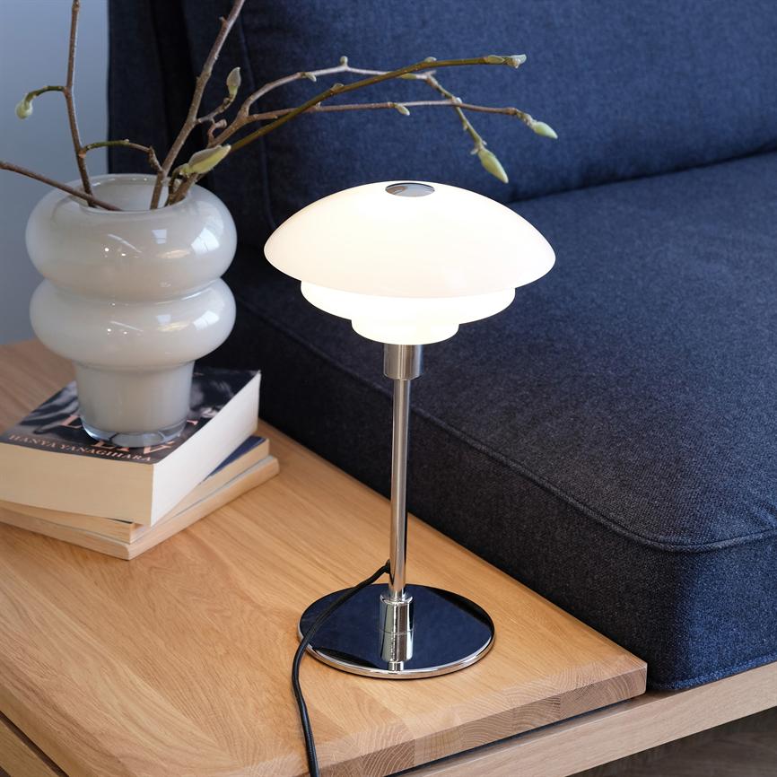 Eine moderne weiße Tischlampe leuchtet sanft auf einem Holztisch; Es steht neben einer weißen Vase mit Knospen und einem Stapel Bücher vor einem dunkelgrauen Sofa im Wohnzimmer.